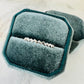 A silver chevron-patterned band ring displayed in an open teal velvet ring box on a light marble surface.