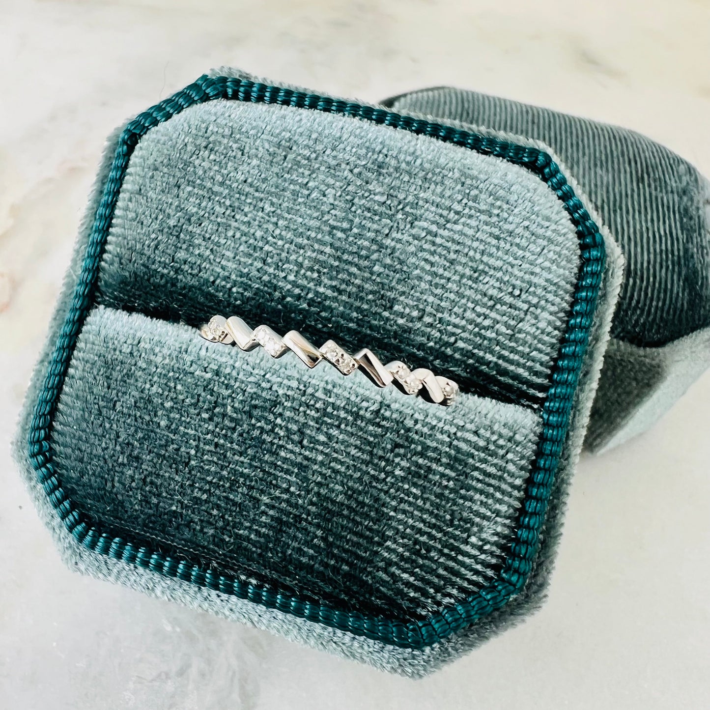 A silver chevron-patterned band ring displayed in an open teal velvet ring box on a light marble surface.