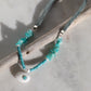 A beaded necklace with turquoise-blue chips, a white seashell charm, and a small turquoise accent, displayed on a driftwood branch over a light marble surface.