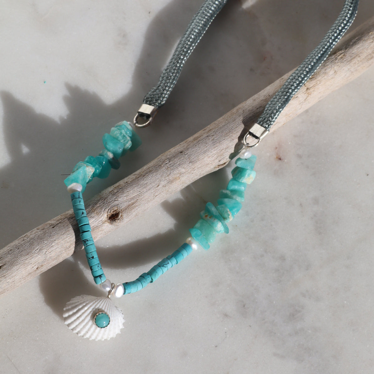 A beaded necklace with turquoise-blue chips, a white seashell charm, and a small turquoise accent, displayed on a driftwood branch over a light marble surface.