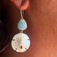 A close-up of a person's ear wearing a silver hoop earring with a light blue teardrop stone and a textured silver sand dollar charm.