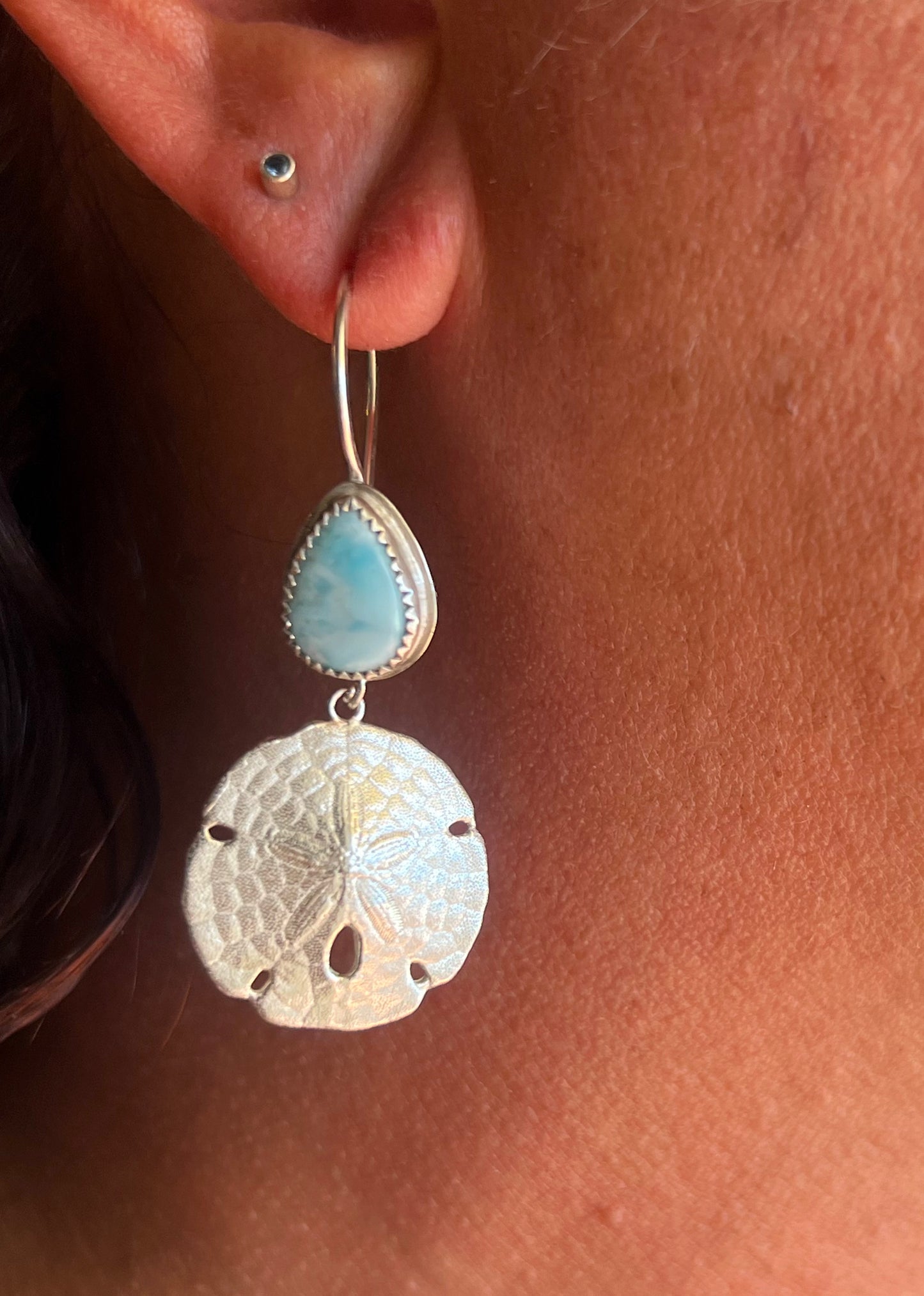 A close-up of a person's ear wearing a silver hoop earring with a light blue teardrop stone and a textured silver sand dollar charm.
