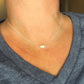 A close-up of a person's neck and upper chest wearing a delicate silver chain necklace with a small, irregularly shaped silver pendant, paired with a blue v-neck t-shirt.