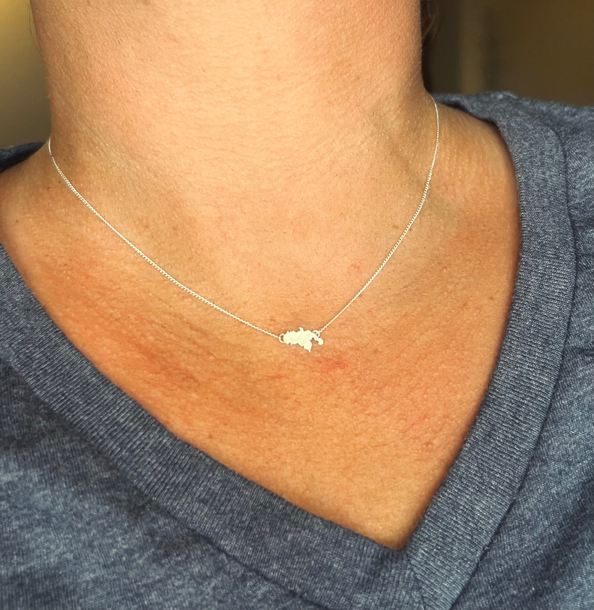 A close-up of a person's neck and upper chest wearing a delicate silver chain necklace with a small, irregularly shaped silver pendant, paired with a blue v-neck t-shirt.