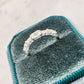 A silver ring with a row of clear stones, displayed on a teal velvet ring box against a light marble background.