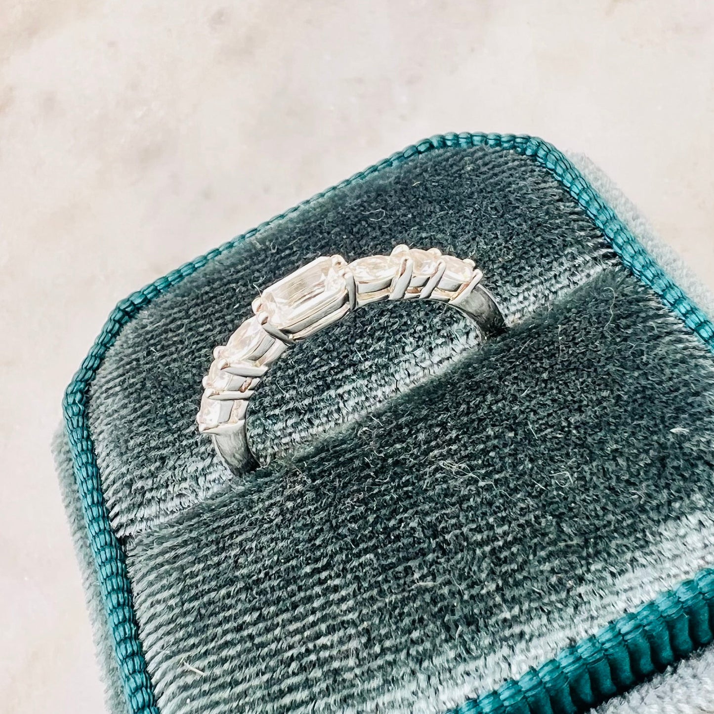 A silver ring with a row of clear stones, displayed on a teal velvet ring box against a light marble background.