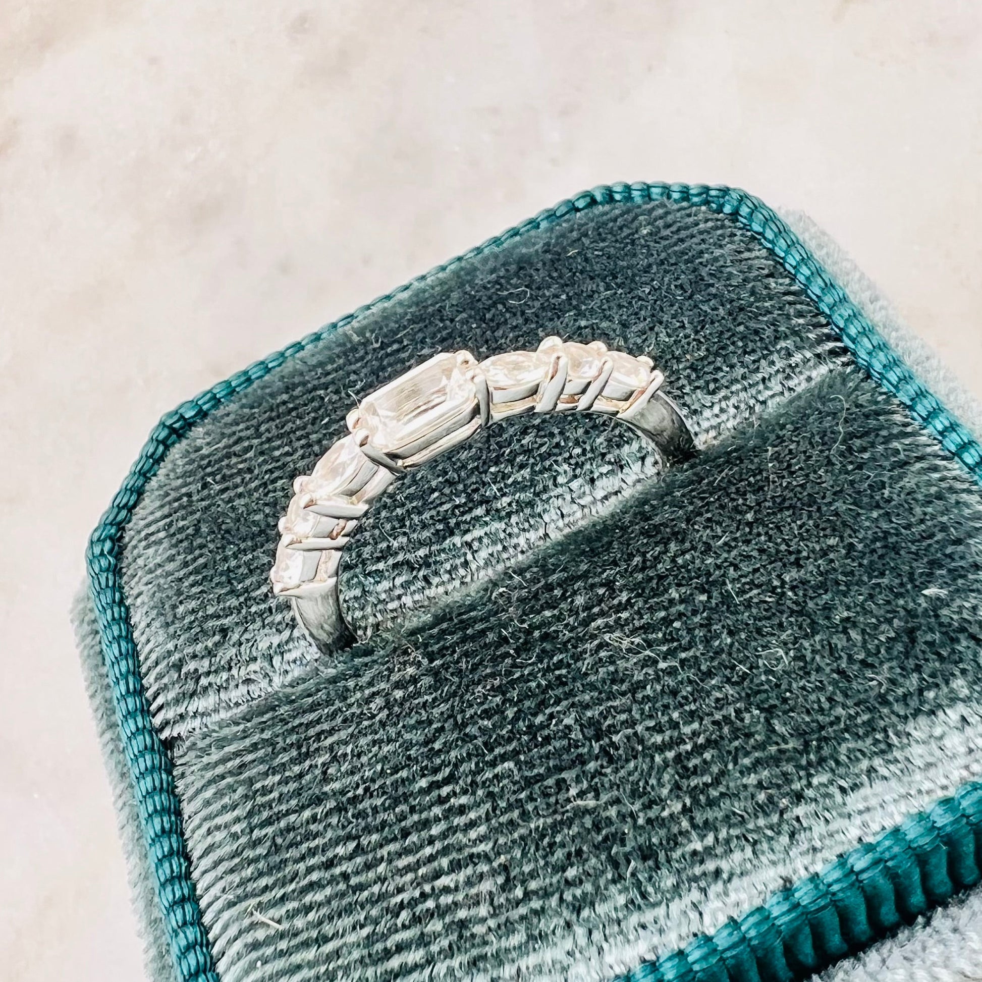 A silver ring with a row of clear stones, displayed on a teal velvet ring box against a light marble background.